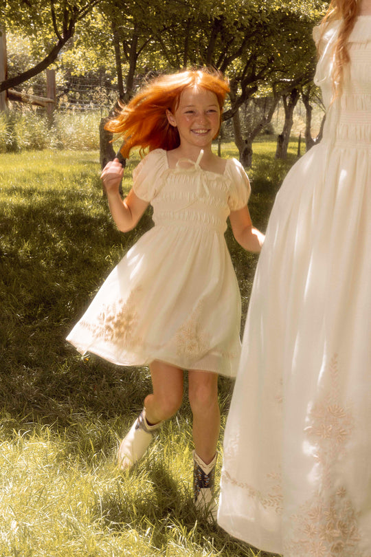 A young girl with reddish hair, wearing the Ivy City Co Mini Mae Dress in Metallic Thread and cowboy boots, smiles as she runs on grass near a woman in a matching dress, both surrounded by sunlit trees.