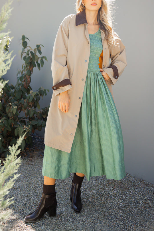 Woman wearing a beige coat over a green dress with black boots, standing against a light gray wall.