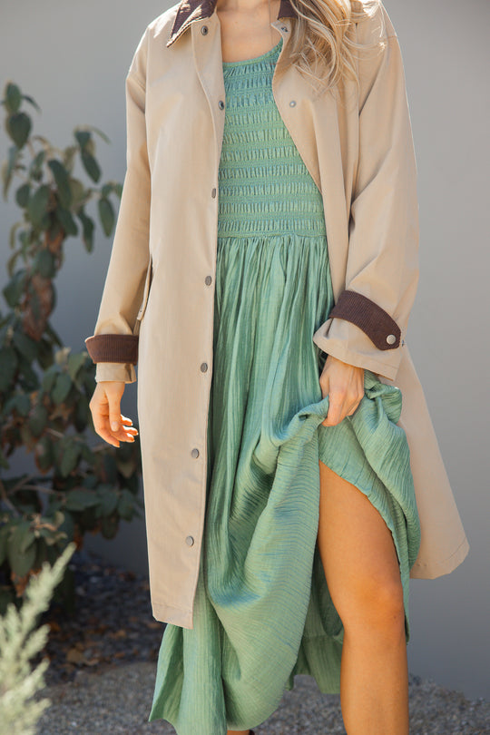 Person wearing a green dress and beige coat with brown trim outdoors.