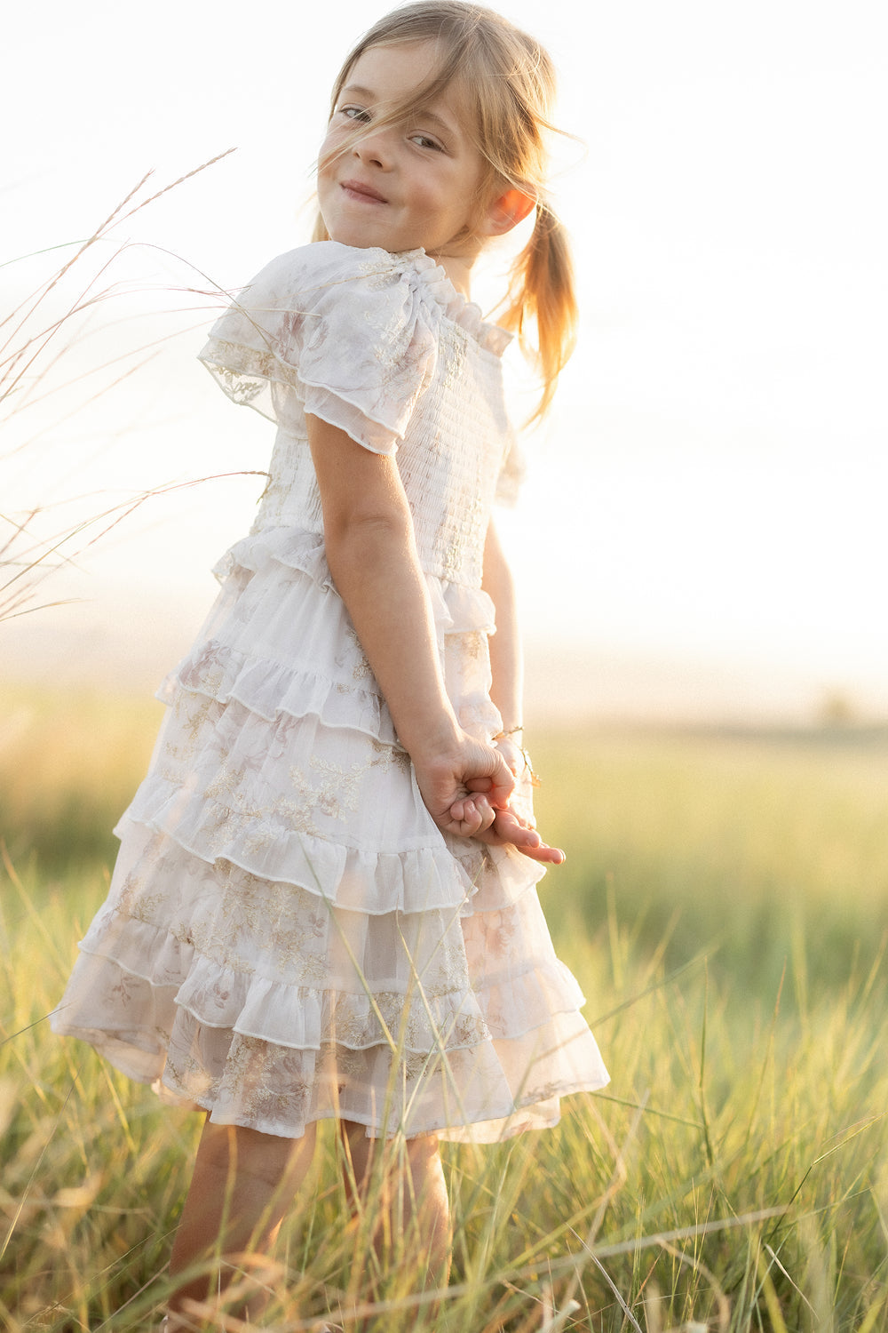 A young girl in the Ivy City Co Mini Winnie Dress in Bright White Wispy Wildflower stands in a grassy field, looking over her shoulder and smiling softly as sunlight illuminates the scene.