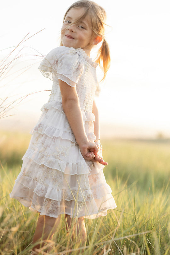 A young girl in the Ivy City Co Mini Winnie Dress in Bright White Wispy Wildflower stands in a grassy field, looking over her shoulder and smiling softly as sunlight illuminates the scene.