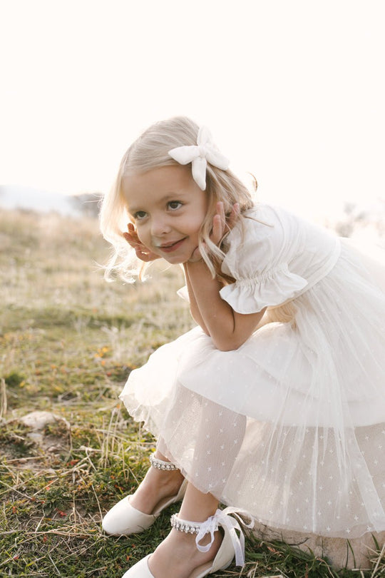 A young girl beams playfully, kneeling on the grass in gentle sunlight while wearing the Ivy City Co Mini Ballerina Dress in White and a matching bow, with her hands held up to her face.