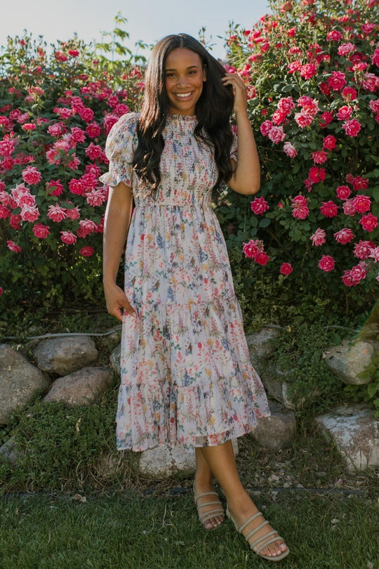 A woman wearing the Ivy City Co Cordelia Floral Dress stands smiling in front of a lush rose bush with pink blooms, one hand touching her hair. She pairs the maternity-friendly floral dress with sandals and is surrounded by greenery and rocks.