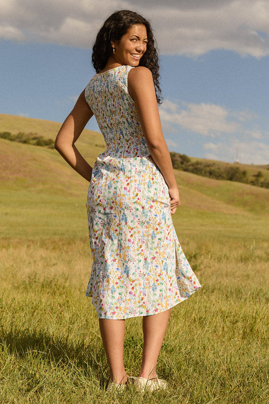 Wearing the Ana Dress in Floral Fields by Ivy City Co, a woman stands on grass, smiling over her shoulder. The sleeveless, maternity-friendly midi dress with a smocked bodice features a delicate floral print. Behind her, green hills and a partly cloudy sky create an idyllic backdrop for this charming look.