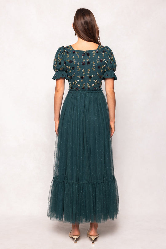 A woman stands with her back to the viewer, wearing Ivy City Co’s Adara Dress in Green—a maxi dress featuring puffed sleeves, intricate floral embroidery on the bodice, and a sheer tulle overlay. Her long brown hair flows down her back, and she completes the look with cream-colored shoes.