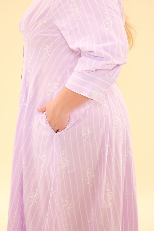 A person stands sideways with one hand in a side pocket, wearing the Cameron Dress in Lavender by Ivy City Co—a long-sleeved, midi shirt dress featuring white stripes and patterns.