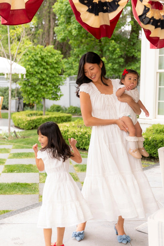 A woman wearing the Ivy City Co Mini Palermo Dress in White smiles while holding a baby dressed in a matching summer dress. Nearby, a young girl dances in another white Mini Palermo Dress. They are outdoors beneath festive red, white, and blue bunting.