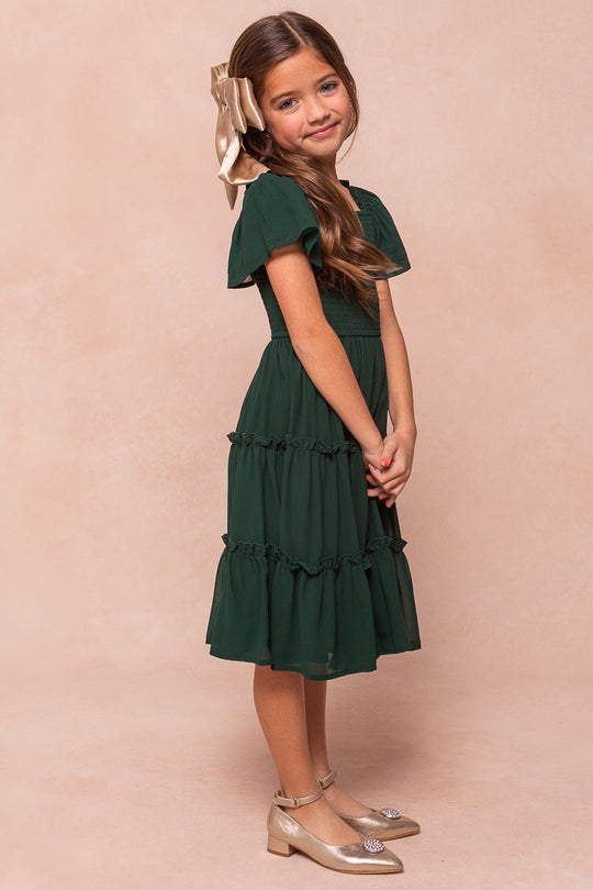 A young girl wears the Ivy City Co Mini Ellie Dress in Emerald Chiffon with ruffled sleeves, a tiered skirt, and smocked bodice, styled with gold flats and a matching hair bow.