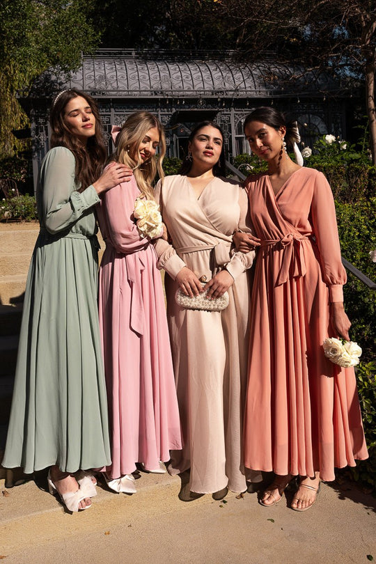 Four women stand outdoors on stone steps in front of a garden gazebo, wearing long, flowing pastel maxi dresses—including the Andie Dress in Apricot Crush by Ivy City Co. They pose together, smiling, holding small bouquets and a clutch.