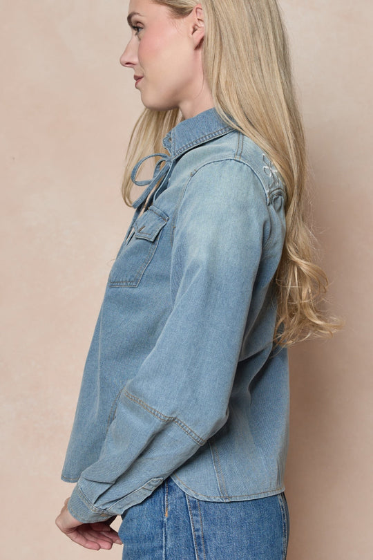 A woman with long blonde hair stands in profile against a beige background, wearing the Ivy City Co Denver Top in Lightwash Denim—a western-inspired piece featuring pearl snap buttons—paired with light blue jeans.
