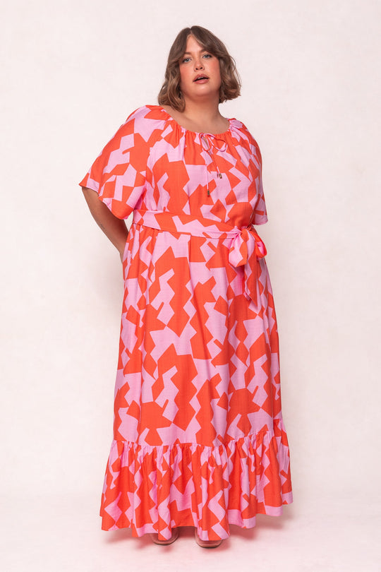A woman wears the Ivy City Co Haven Dress in Geometric—a pink and orange maxi dress featuring a geometric pattern, short sleeves, ruffled hem, and a matching belt—as she stands against a plain light background.