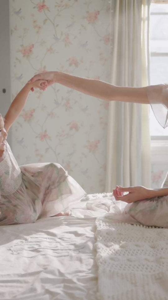A child and an adult sit on a bed in a softly lit room decorated with floral wallpaper, gently holding hands and wearing light, flowing dresses. The adult is dressed in the Rhiannon Dress in Painted Bloom by Ivy City Co—a gorgeous, maternity friendly dress that’s perfect for sharing special moments together. Warm sunlight streams through a nearby window.