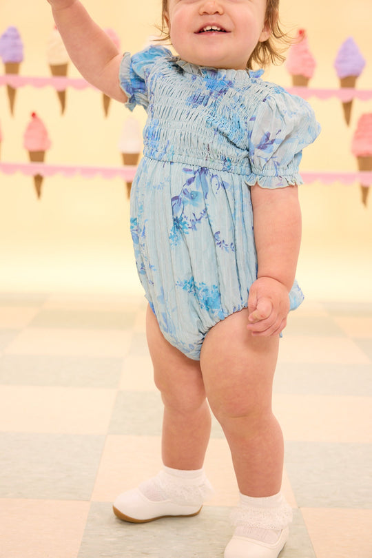 A toddler wearing the Ivy City Co Baby Flynn Romper in Aquarelle Fleur and white shoes stands on a checkered floor, with ice cream cone decorations hanging in the blurred background.