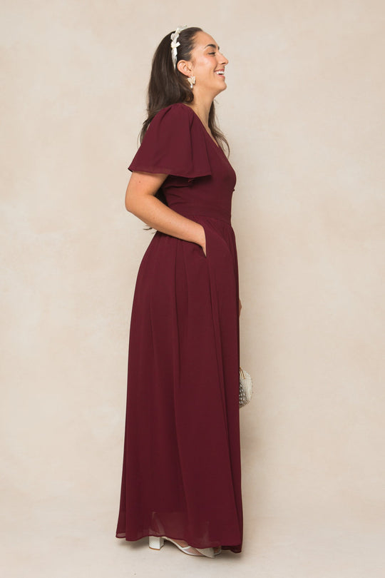 A woman with long dark hair, white earrings, and a headband stands sideways, smiling in the Abbie Dress in Wine Chiffon by Ivy City Co. The floor-length dress features short flutter sleeves and is paired with a small beaded clutch. Perfect for fall occasions or as a bridesmaid dress, she stands out against the beige background.