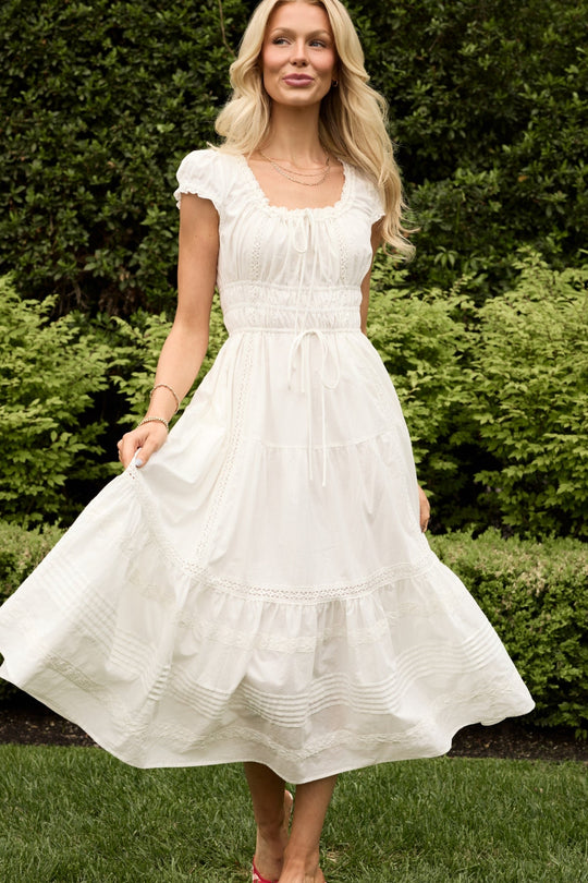 A woman wearing the Palermo Dress in White from Ivy City Co stands outdoors, holding the skirt of her white, ruffled, short-sleeve dress. She has long blonde hair and is smiling, with green bushes and trees in the background. This elegant style is both nursing friendly and maternity friendly.