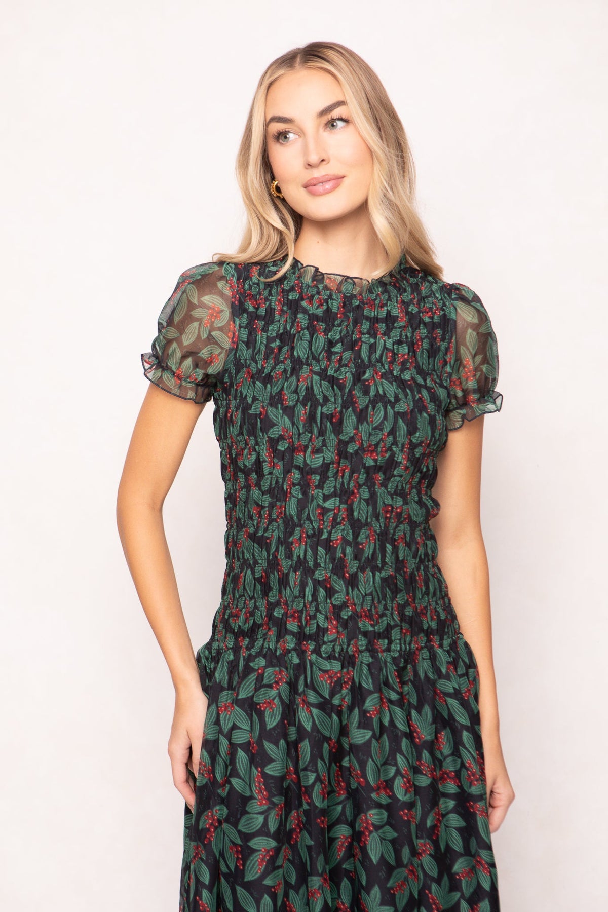 A woman wears a black, maternity-friendly Hayden Dress in Berries with a smocked bodice, featuring a green leaf and red berry print.
