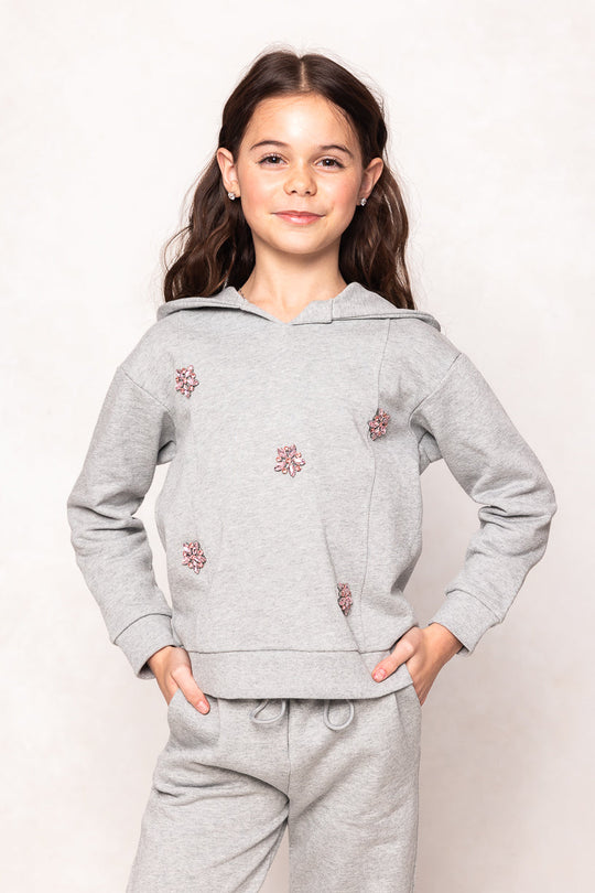 A young girl smiles, wearing the Ivy City Co Mini Skylar Hoodie in Heather Gray with matching sweatpants, both adorned with delicate crystal flower details. She stands with one hand on her hip against a neutral background.