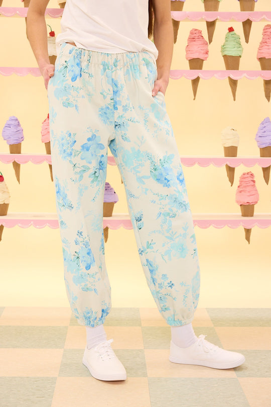 A person wearing Ivy City Co’s Kian Jogger Sweatpants in Blue Aquarelle Fleur, paired with white socks and white sneakers, stands with hands in pockets. Pastel ice cream cones decorate the pink and yellow wall in the background.
