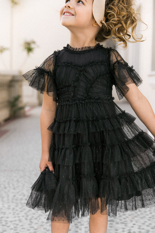 A smiling young girl with curly hair stands outdoors, looking upward, wearing the Ivy City Co Mini Whimsical Dress in Black—a playful style featuring a layered tulle skirt—paired with a beige headband.