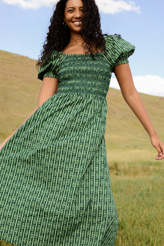 A smiling woman stands in a grassy field with rolling hills and a blue sky behind her, enjoying the day in the Ivy City Co Sunday Best Dress in Pine Felicity Floral—a beautiful outfit designed to be both nursing- and maternity-friendly.