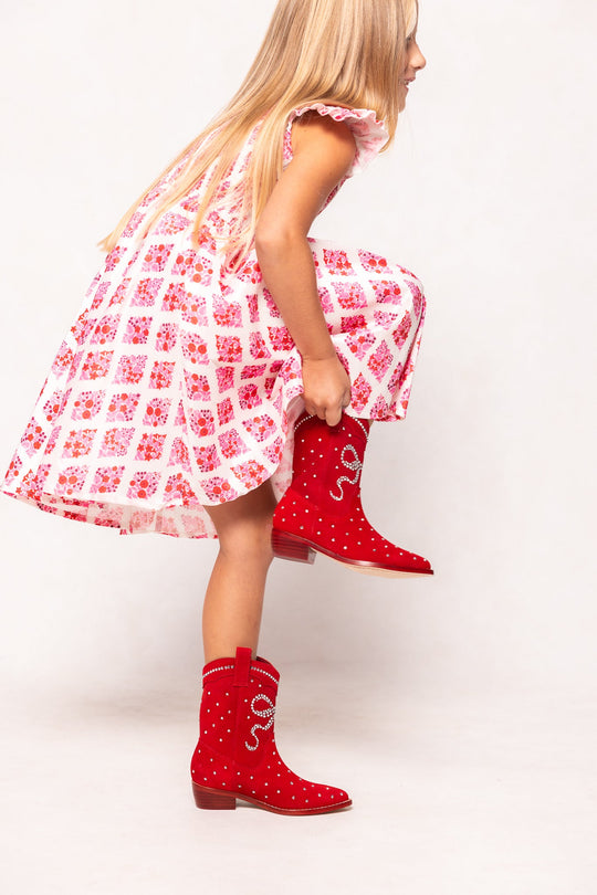 A young girl wearing a pink and white floral dress bends down to slip on the Ivy City Co Mini Channing Bootie in Red—bright, studded cowboy boots featuring a sparkly snake design that are perfect for matching mommy-and-me looks.