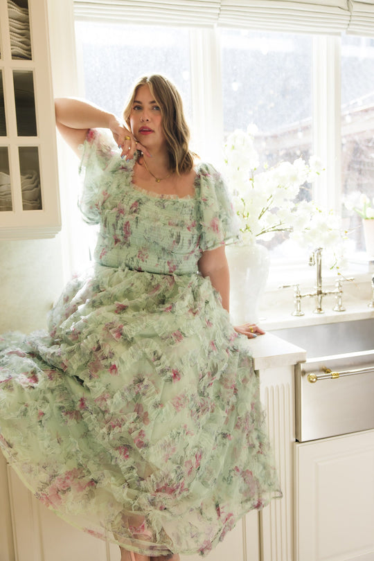 A woman wearing the Florentina Dress in Pistachio Aquarelle Fleur by Ivy City Co stands by a sunlit kitchen sink, surrounded by white cabinets, a large window, and a vase of white flowers. This elegant, maternity-friendly dress combines comfort and style for everyday moments.