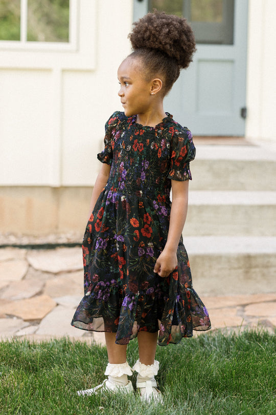 A young girl with curly hair in a ponytail wears the Ivy City Co Mini Cordelia Dress in Evergreen Vivid Meadow, which features puff sleeves, along with white ruffled shoes. She stands on grass, looking to her left, in front of a house with stone steps.
