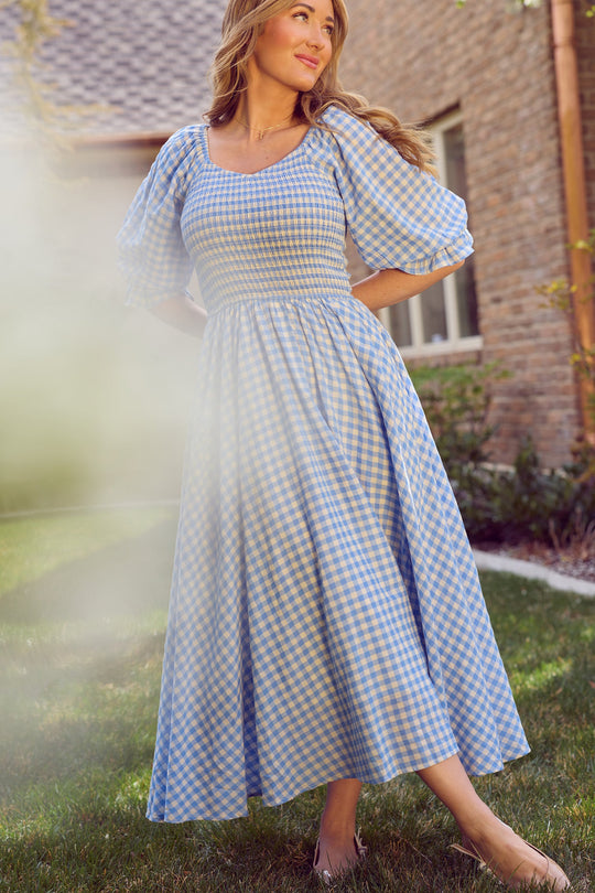 A woman wearing the Ivy City Co Teagan Dress in Blue Gingham—with puff sleeves and nursing-friendly design—stands on the grass, smiling and looking to the side, with a brick building behind her. This stylish dress is perfect for both maternity wear and nursing, ensuring comfort without compromising on charm.