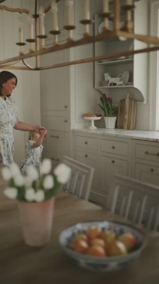 A woman wearing the Ivy City Co Sophia Dress in Blue Bouquet holds a child’s hands in a bright kitchen, where tulips and fruit adorn the counter and table, creating a cozy and welcoming atmosphere.