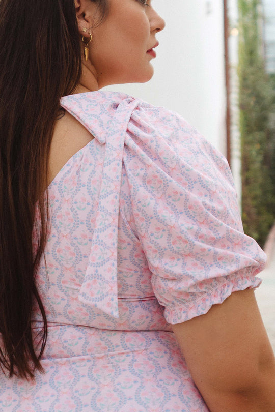 A woman with long brown hair wears the Ivy City Co Sailor One Piece in Raquel Pink Floral, featuring puff sleeves, an asymmetrical neckline, and a shoulder bow. She stands outdoors, turned slightly to the side, wearing gold hoop earrings.