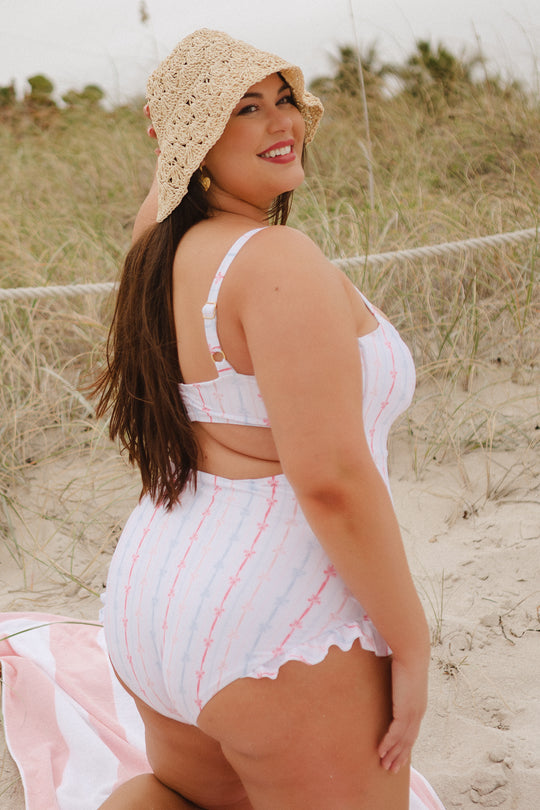 A woman kneels on the sand at the beach, smiling over her shoulder in the Ivy City Co Lucy One Piece in Breea Bow and a straw sunhat. Grassy dunes and a towel appear in the background.