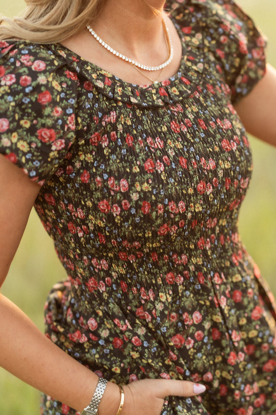 Wearing the Annalise Dress in Obsidian Vintage Poppies by Ivy City Co, paired with a pearl necklace, watch, and bracelets, a person stands outdoors in warm lighting with one hand in their pocket.