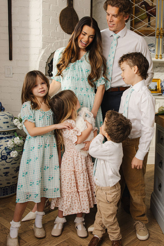 A smiling family of five, dressed in coordinated pastel outfits, stands together in a kitchen. The children gather around a young girl wearing the Ivy City Co Mini Jessie Dress in Bloom Gingham as she holds a white rabbit, while the parents look on happily.