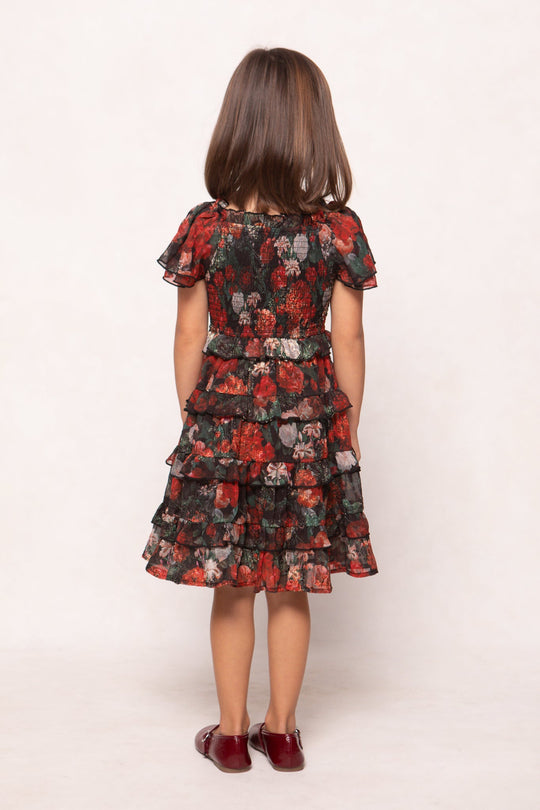 A young girl with shoulder-length brown hair stands facing away, wearing the Ivy City Co Mini Winnie Dress in Midnight Roses—a ruffled, floral-patterned dress in dark red, green, and beige—paired with matching red shoes, set against a plain white background.