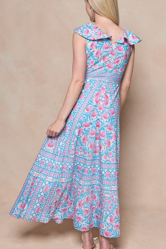 A woman with long blonde hair is seen from behind, wearing the Ivy City Co Samara Dress in Thistle Flower Mosaic Florise—a blue and pink sleeveless dress featuring a bow at the back.