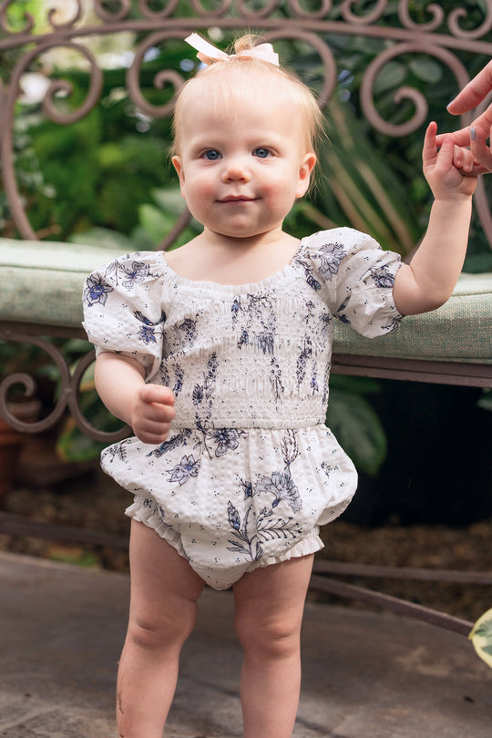 A toddler with light hair and blue eyes, dressed in the Ivy City Co Baby Roselyn Romper in Blue Enchanted Floral featuring charming puff sleeves, stands holding an adult’s hand. Green plants and a decorative bench appear in the background.