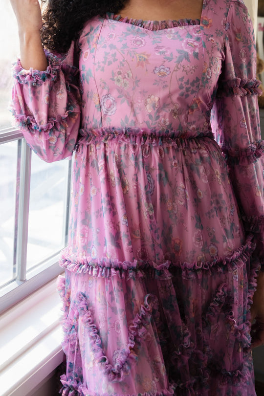 A person stands by a window wearing the Ivy City Co Catherine Dress in Climbing Vine Bloom, featuring long sleeves and delicate ruffle details. The image highlights the dress from the shoulders to the knees.