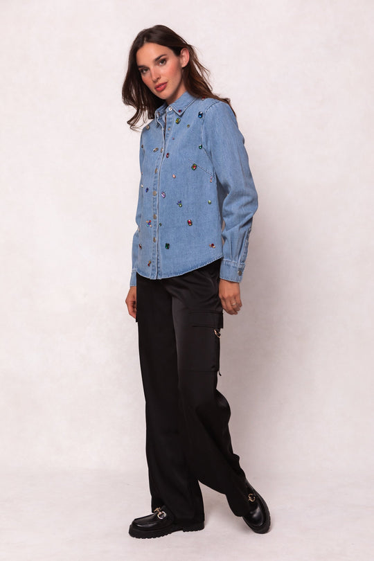 A woman stands against a plain light background, posing with one leg slightly forward and her hand in her pocket, wearing a light blue, jewel-embellished denim shirt paired with the Shay Satin Pants in Black by Ivy City Co—featuring a loose fit and elastic waistband—and black loafers.