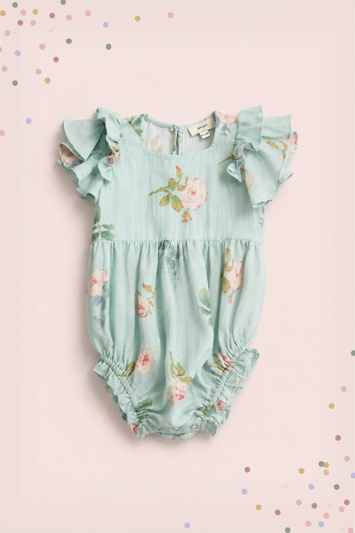 The Baby Hattie Romper in Roselyn Blue by Ivy City Co is a light blue romper featuring pink floral prints and ruffled sleeves, beautifully displayed on a soft pink background adorned with colorful confetti dots in the corners. This charming piece is perfect for making any mommy-and-me outfit stand out.