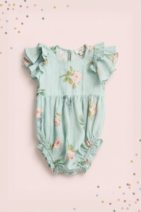 The Baby Hattie Romper in Roselyn Blue by Ivy City Co is a light blue romper featuring pink floral prints and ruffled sleeves, beautifully displayed on a soft pink background adorned with colorful confetti dots in the corners. This charming piece is perfect for making any mommy-and-me outfit stand out.