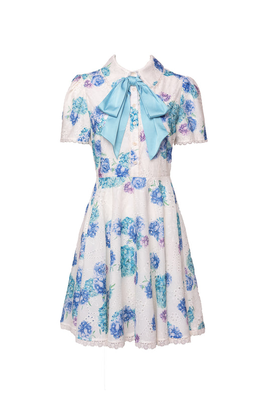The Penny Dress in Blue Floral by Ivy City Co features short sleeves, a collared neckline with a light blue bow, white base with blue and purple floral print, lace trim, fitted waist, and flared knee-length skirt for a charming look.