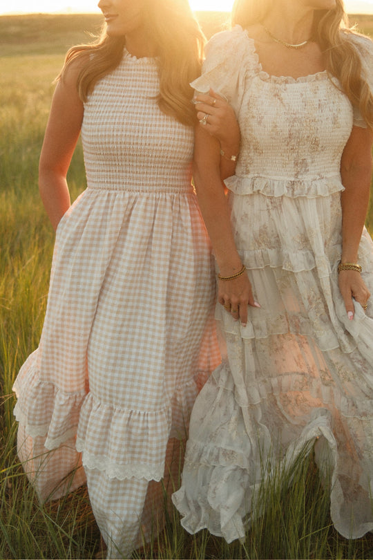 Two women stand arm in arm in a sunlit field, wearing flowy, light-toned dresses with ruffles and smocked bodices. One of them wears the Presley Sleeveless Dress in Cream Gingham by Ivy City Co—a feminine, maternity-friendly piece. Only their bodies are shown from the neck down.