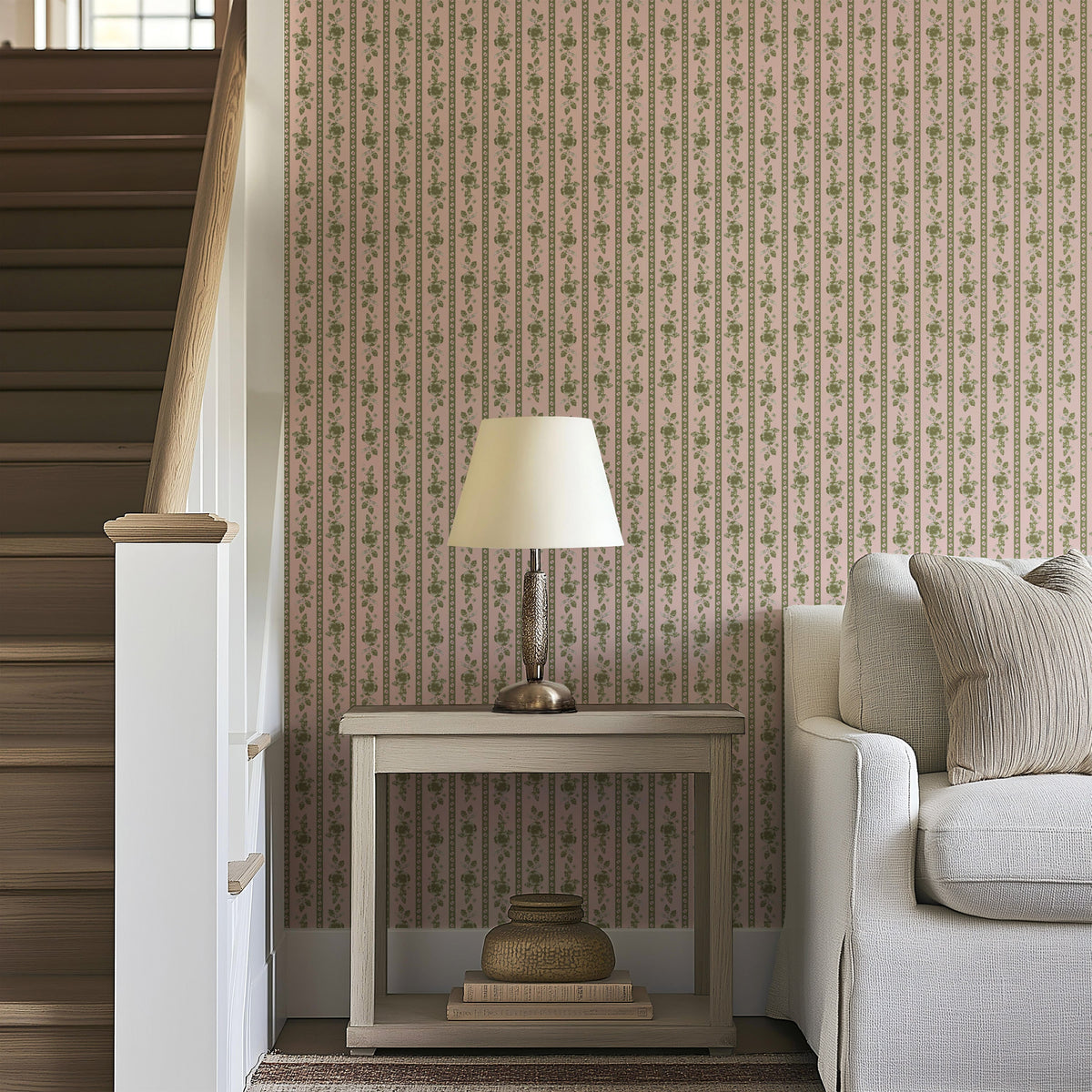 A side table with a lamp and stacked baskets sits next to a beige sofa by a staircase, against a wall adorned with Felicity Floral Wallpaper by Loomwell Home Goods in muted tones.