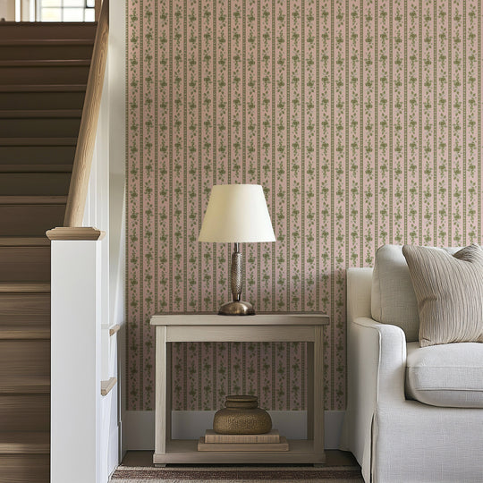 A side table with a lamp and stacked baskets sits next to a beige sofa by a staircase, against a wall adorned with Felicity Floral Wallpaper by Loomwell Home Goods in muted tones.