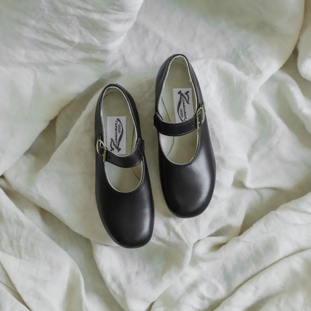 A pair of Mini Mary Janes in Black by Zimmerman Shoes, featuring adjustable ankle straps and crafted from black leather, are displayed side by side on a soft, white, wrinkled fabric background.
