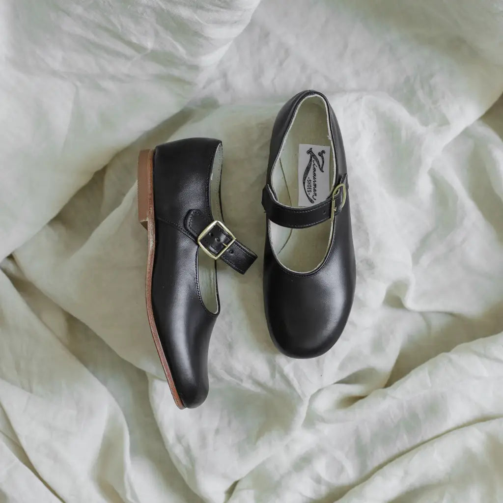 A pair of Zimmerman Shoes Mini Mary Janes in Black, featuring buckle straps and an adjustable ankle strap, rests on a soft, white, crumpled fabric background.
