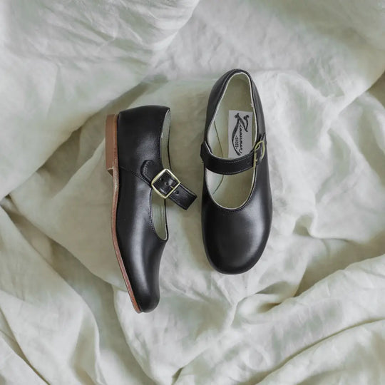 A pair of Zimmerman Shoes Mini Mary Janes in Black, featuring buckle straps and an adjustable ankle strap, rests on a soft, white, crumpled fabric background.