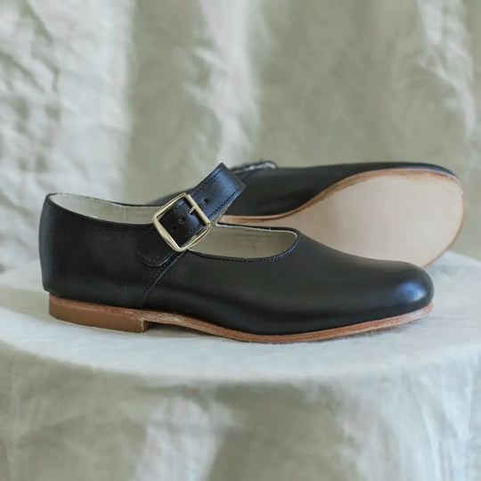 Two Mini Mary Janes in Black by Zimmerman Shoes, crafted from black leather with tan soles and silver buckles, are displayed on a light textured surface—one standing upright and the other laid flat in the background. These shoes for kids feature an adjustable ankle strap to ensure a secure and comfortable fit.