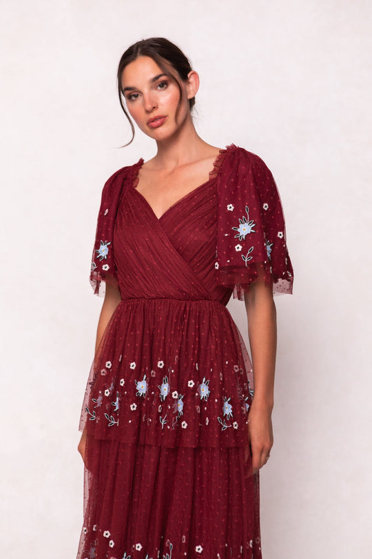 A woman wearing the Mabel Dress in Burgundy by Ivy City Co, featuring short sheer sleeves and blue floral embroidery, stands against a light background.