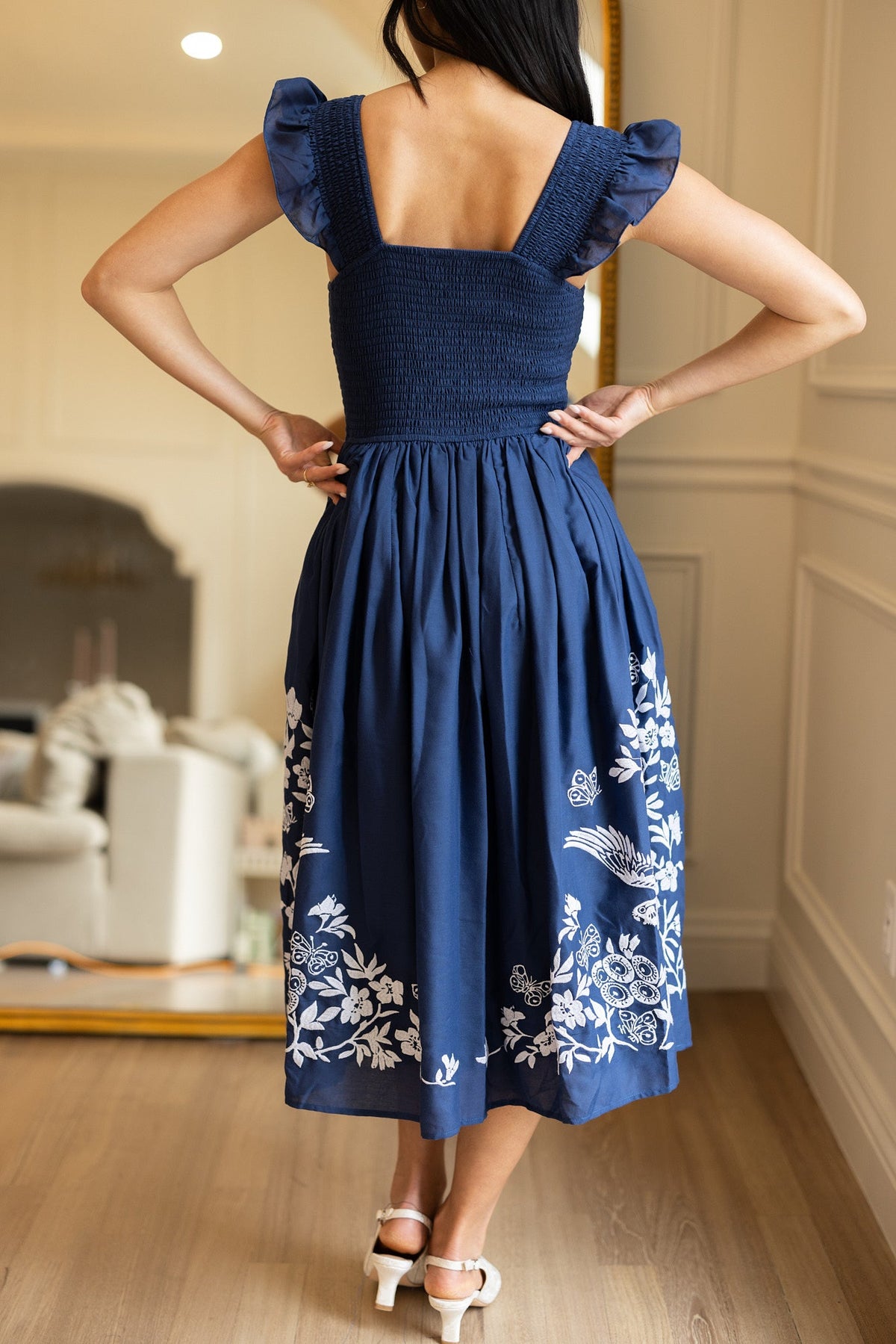 A woman stands indoors, facing away, wearing the Ivy City Co Finch Dress in Blue—a deep blue cotton dress featuring white embroidery on the skirt and ruffled sleeves—paired with white heels. The room has light walls, wood floors, and a mirror reflecting furniture.